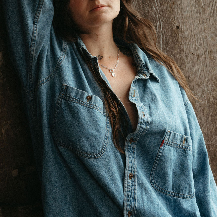 Person wearing a light blue denim shirt against a wooden background wearing the  lupine pendant. 
