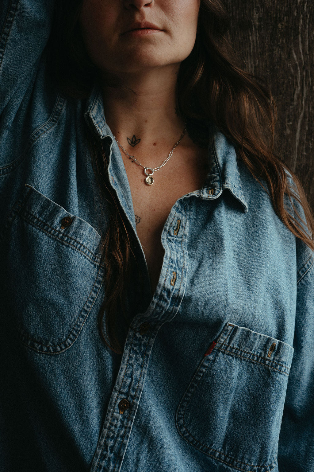 Person wearing a denim shirt with a necklace, against a dark background