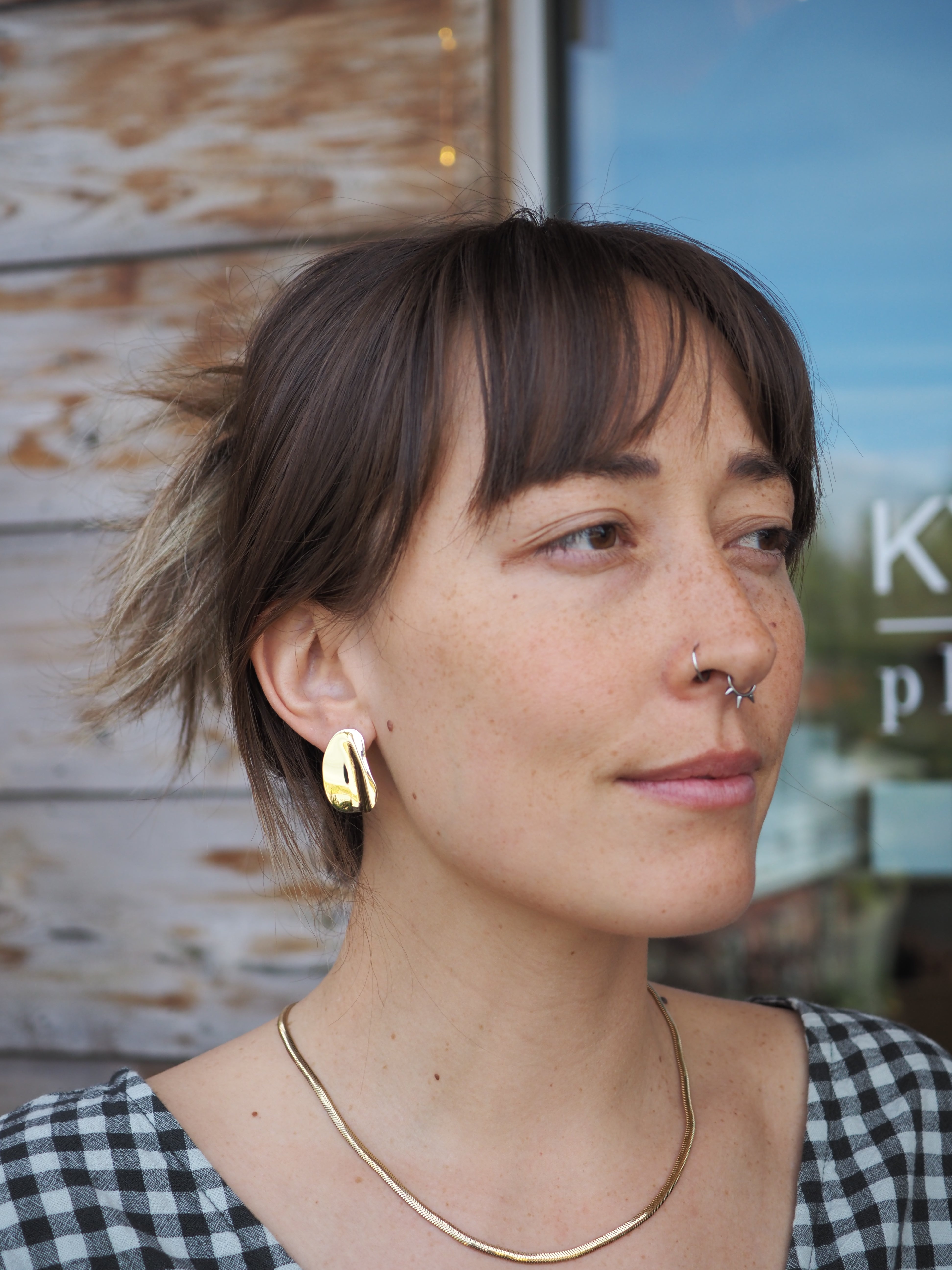 Woman with a nose ring and gold earring and necklace, wearing a checkered top, standing outdoors.