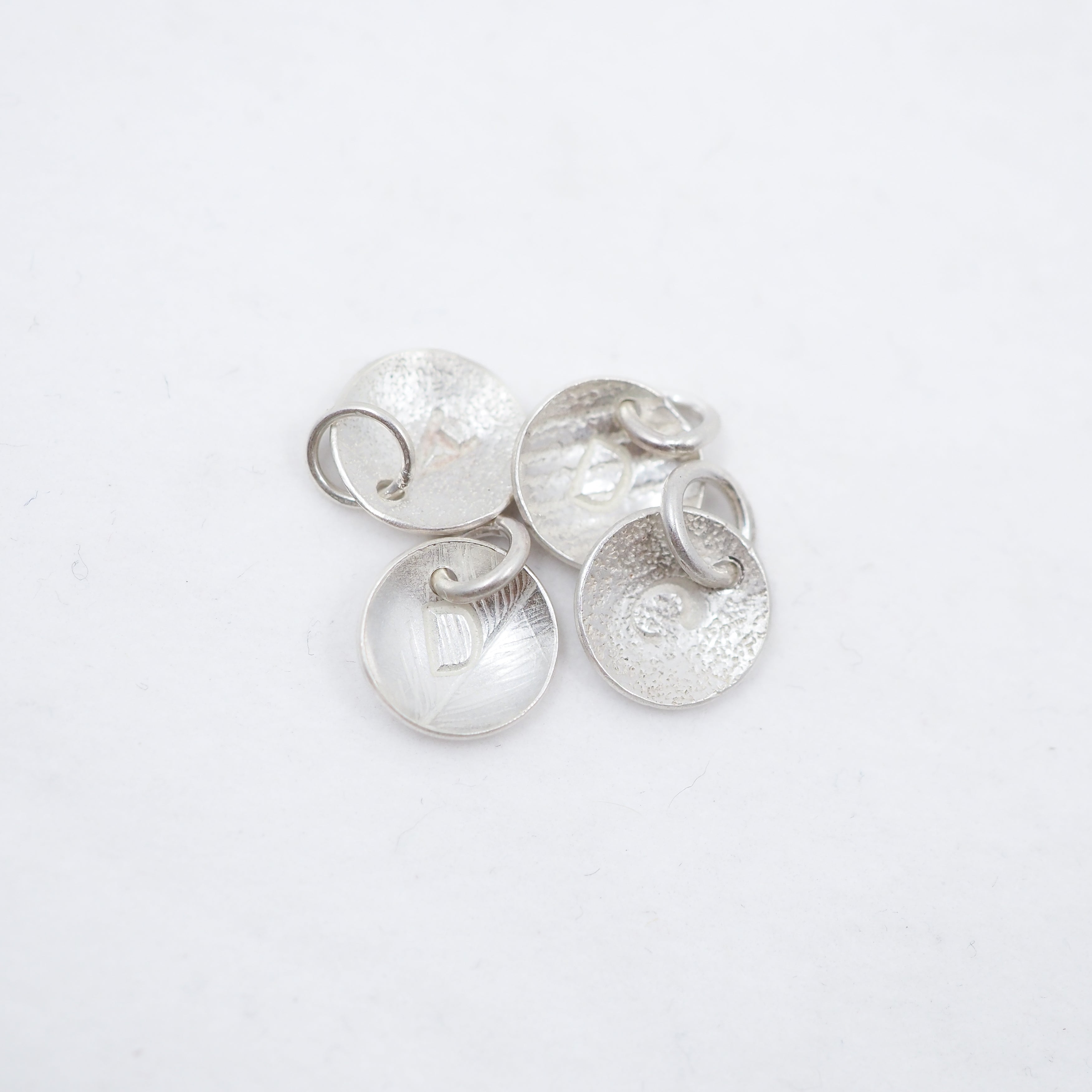 Four textured silver disc charms with attached jump rings, displayed on a clean white background.