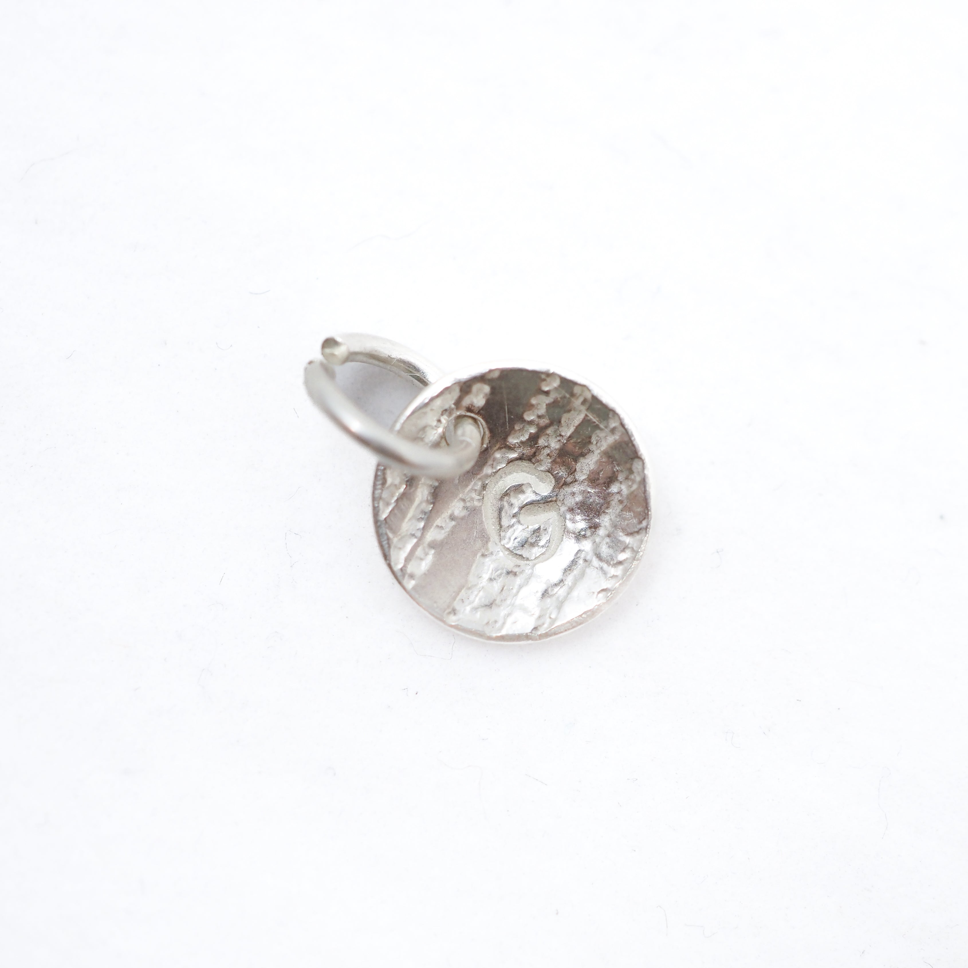 A small silver circular disc charm with a textured, hammered surface and a small attached metal loop, shown on a plain white background.