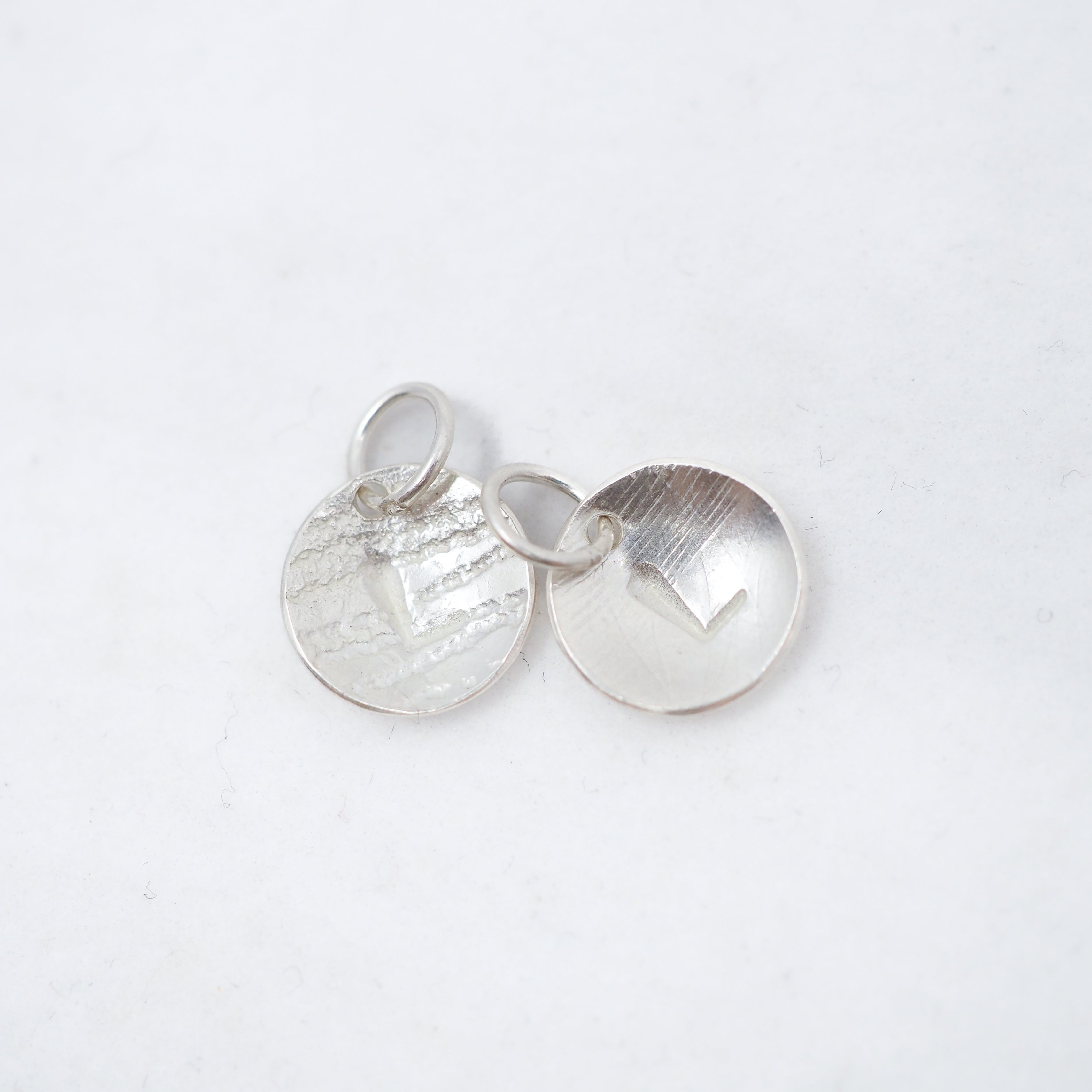 Two small round silver disc charms with textured and brushed finishes, one featuring a raised chevron and the other a heart cutout, shown on a white background.