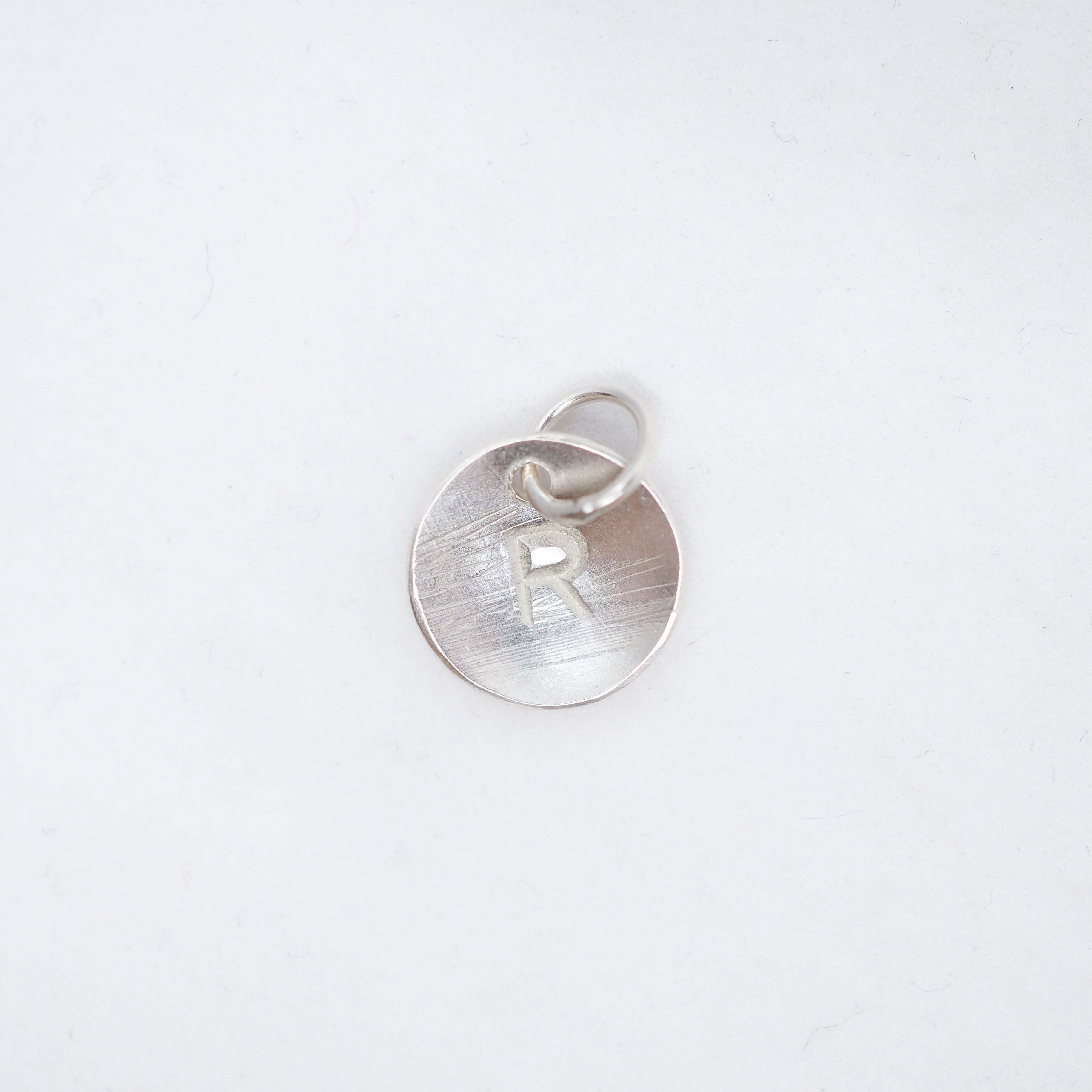 A sterling silver round disc pendant with a brushed finish, featuring a cut-out letter 'R' and a small jump ring at the top, shown on a clean white background.