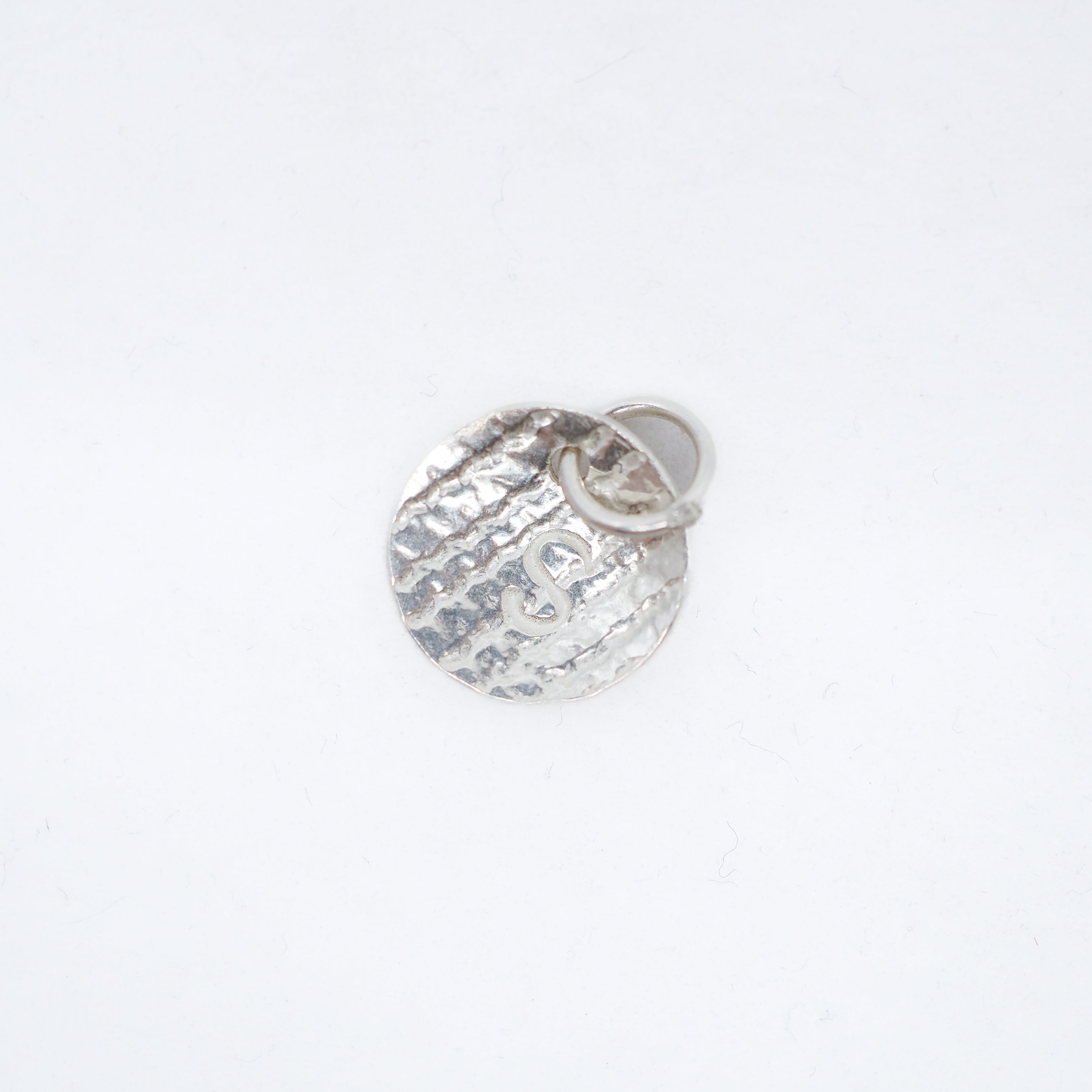 A small hammered silver charm shaped like a round medallion with a small attached loop, photographed on a plain white background.