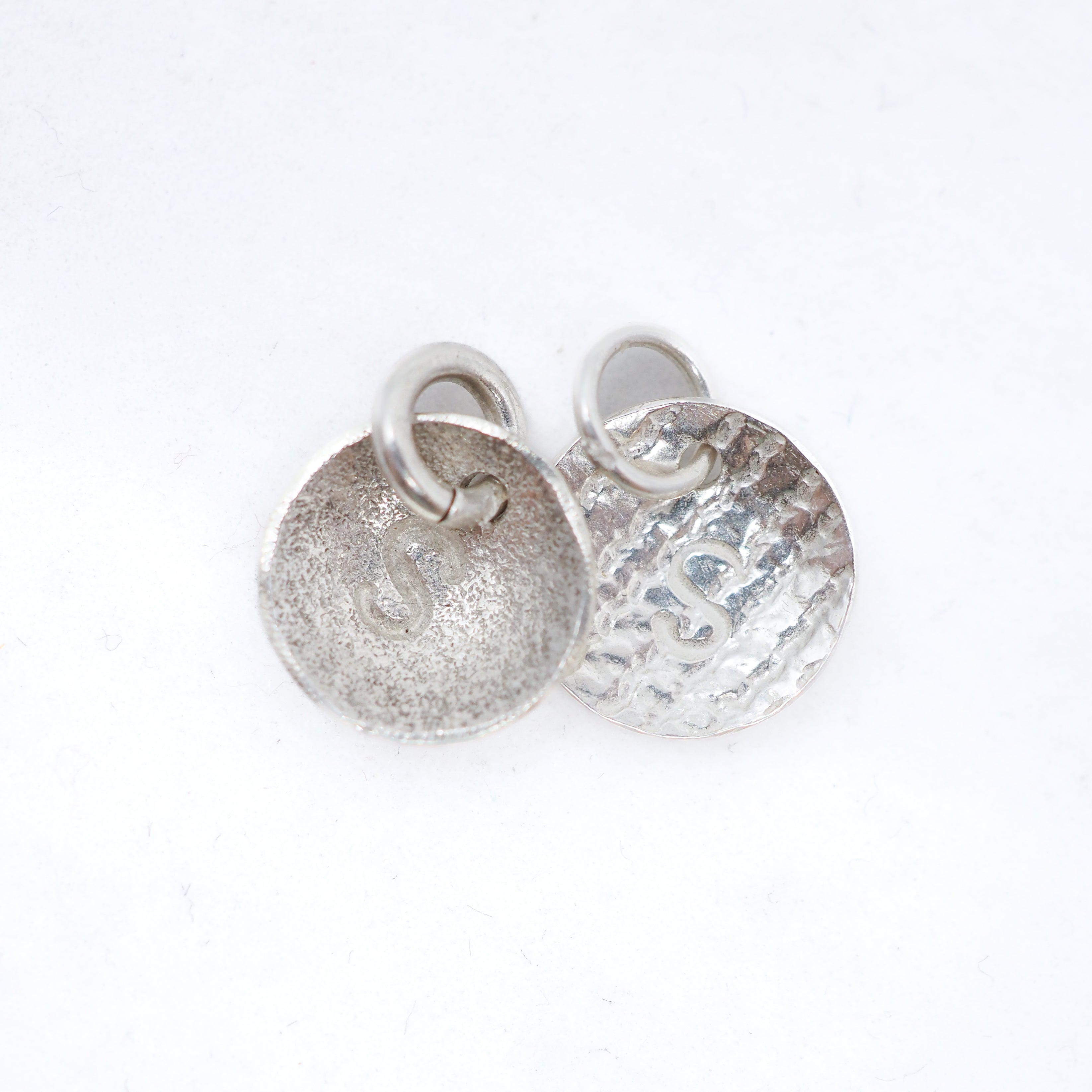 Two silver round disc charms with jump rings, one with a hammered texture and one with a textured matte finish, on a white background.