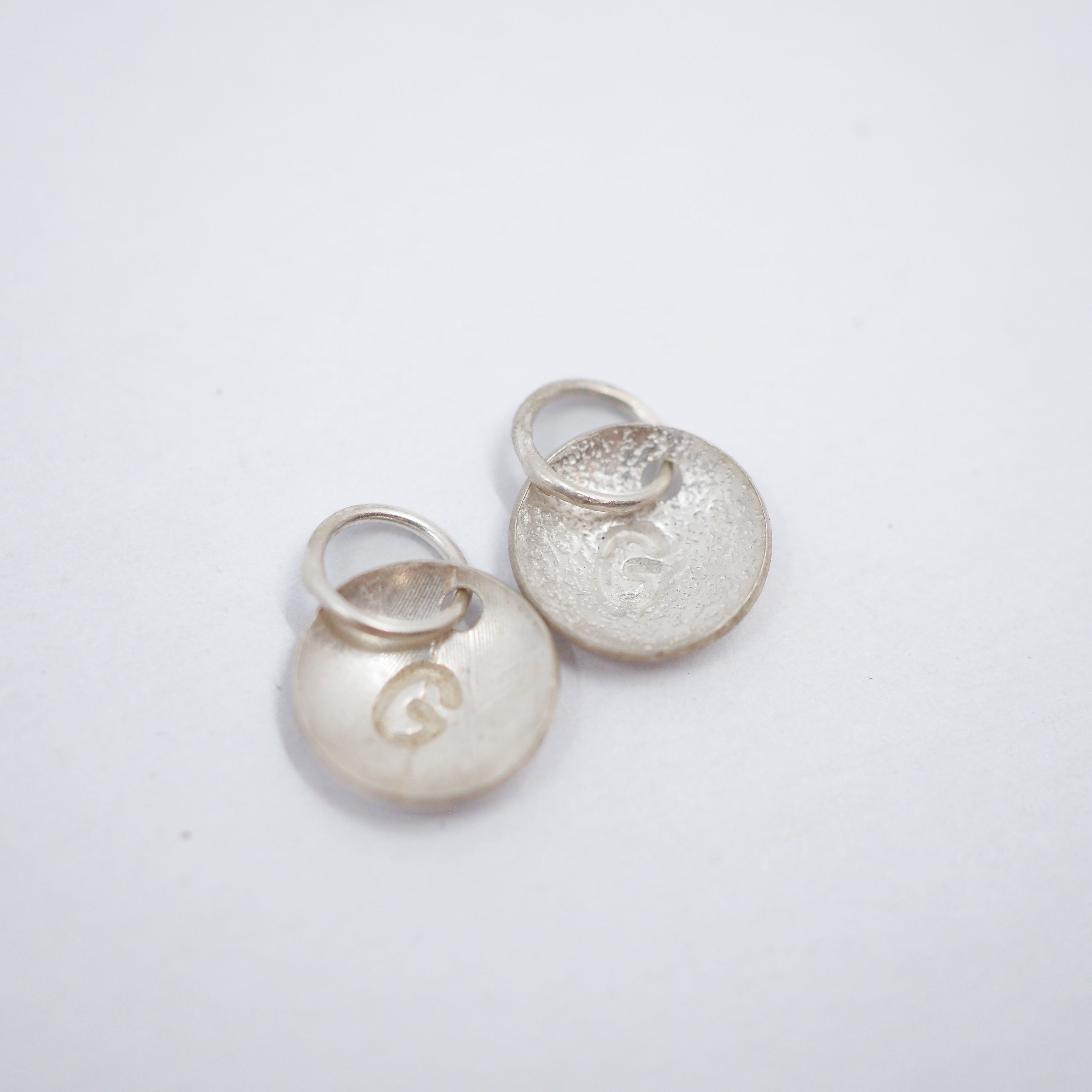 Two small round silver disc charms with attached jump rings, one smooth and one textured, both featuring an engraved letter 'G', on a plain white background.