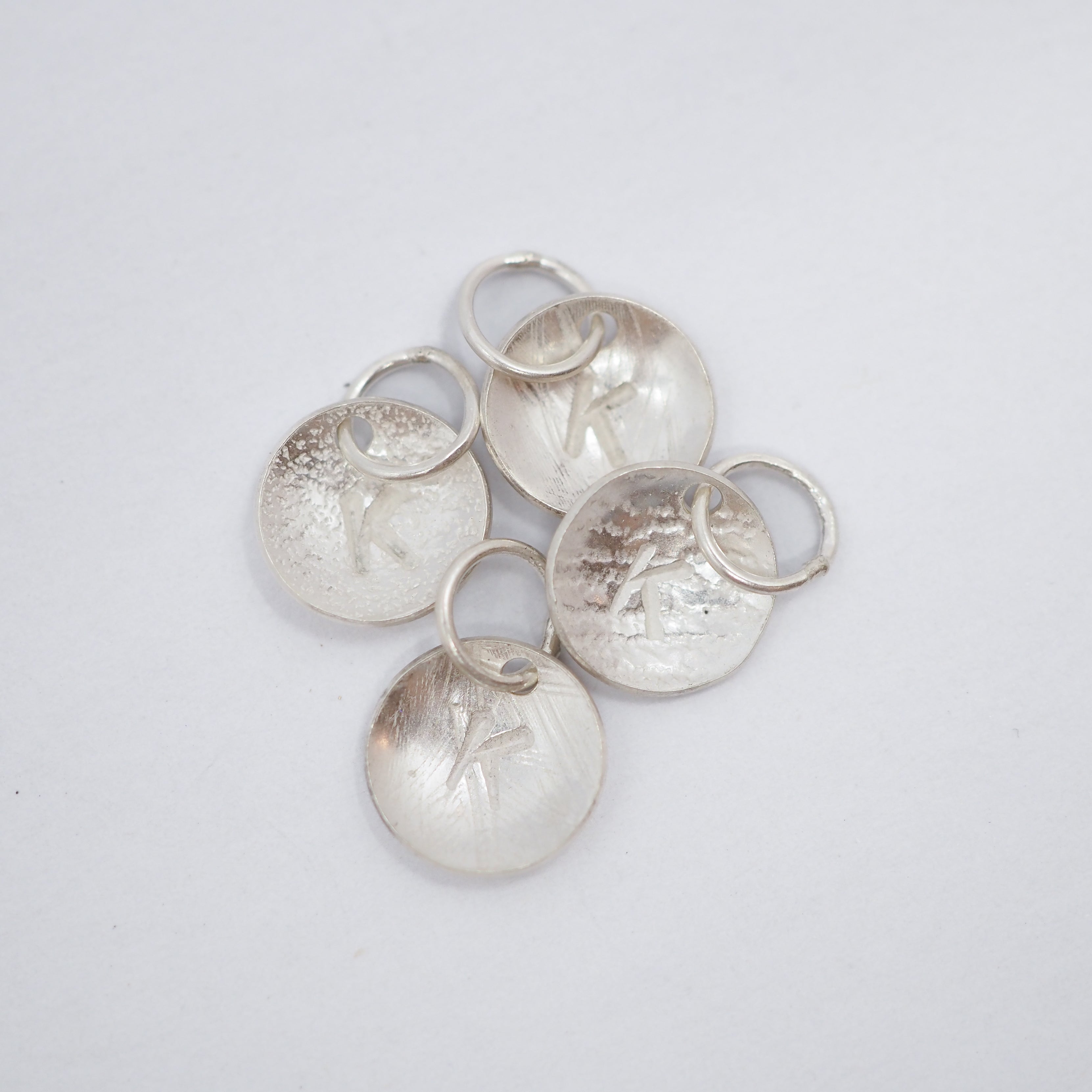 Five silver disk charms with textured finishes and small attached split rings, arranged on a plain light gray background.