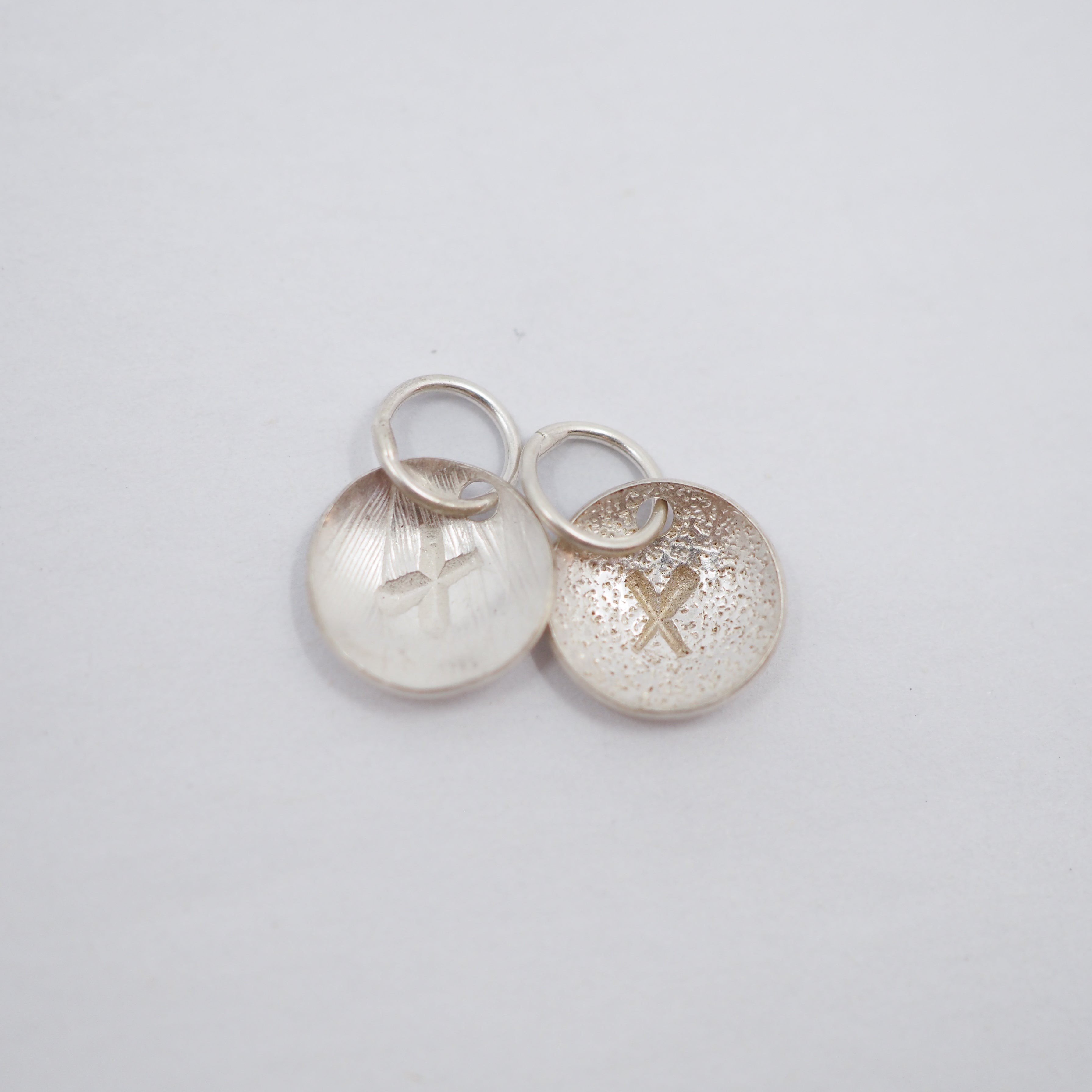 Two small silver oval charms with jump rings, one brushed with a cross and the other textured with a cross, on a light gray background.