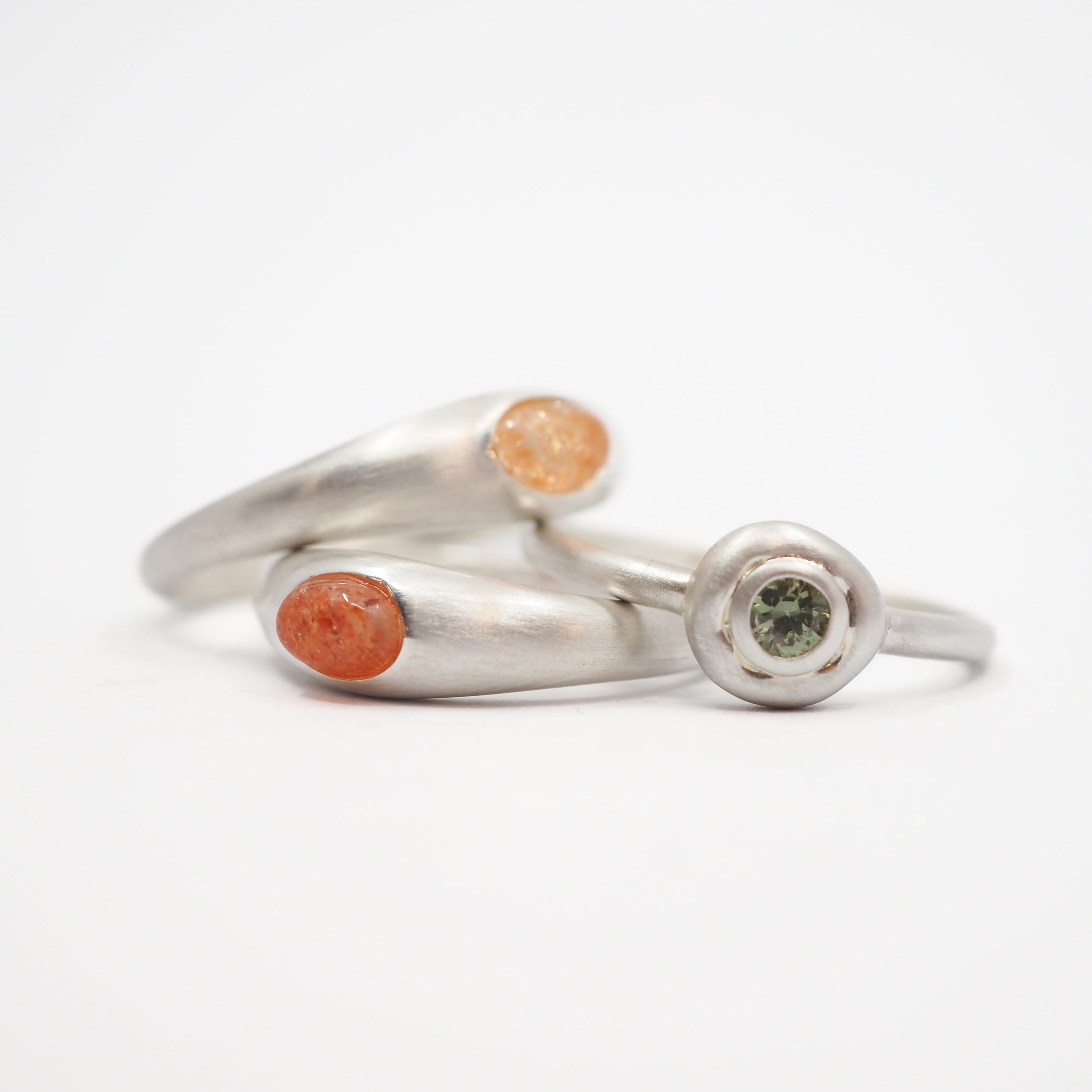 A silver ring with two curved bands, one featuring a round orange stone and the other a bezel-set green stone, photographed on a light gray background.