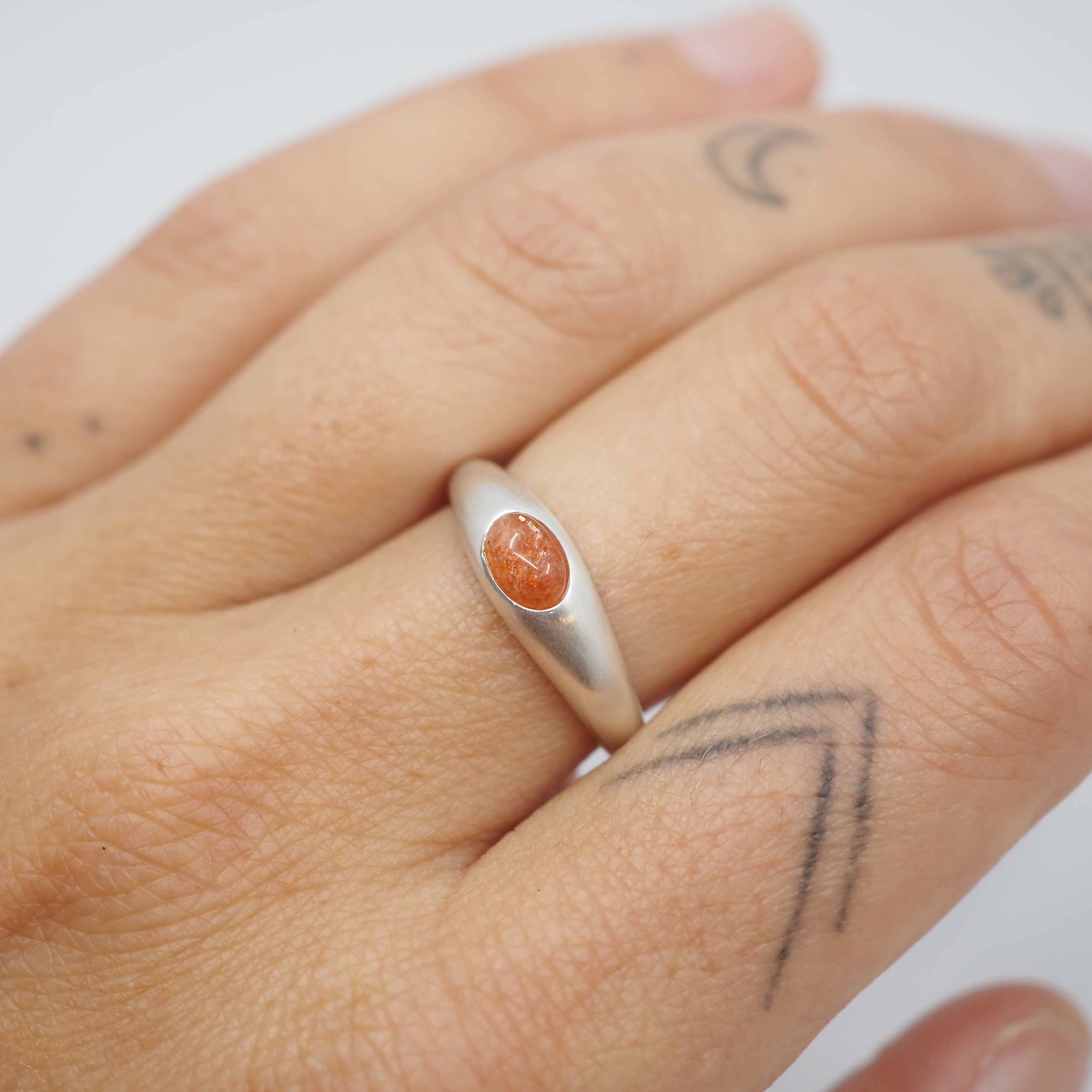A close-up of a hand wearing a silver ring with a small oval orange stone, set against a plain light background.