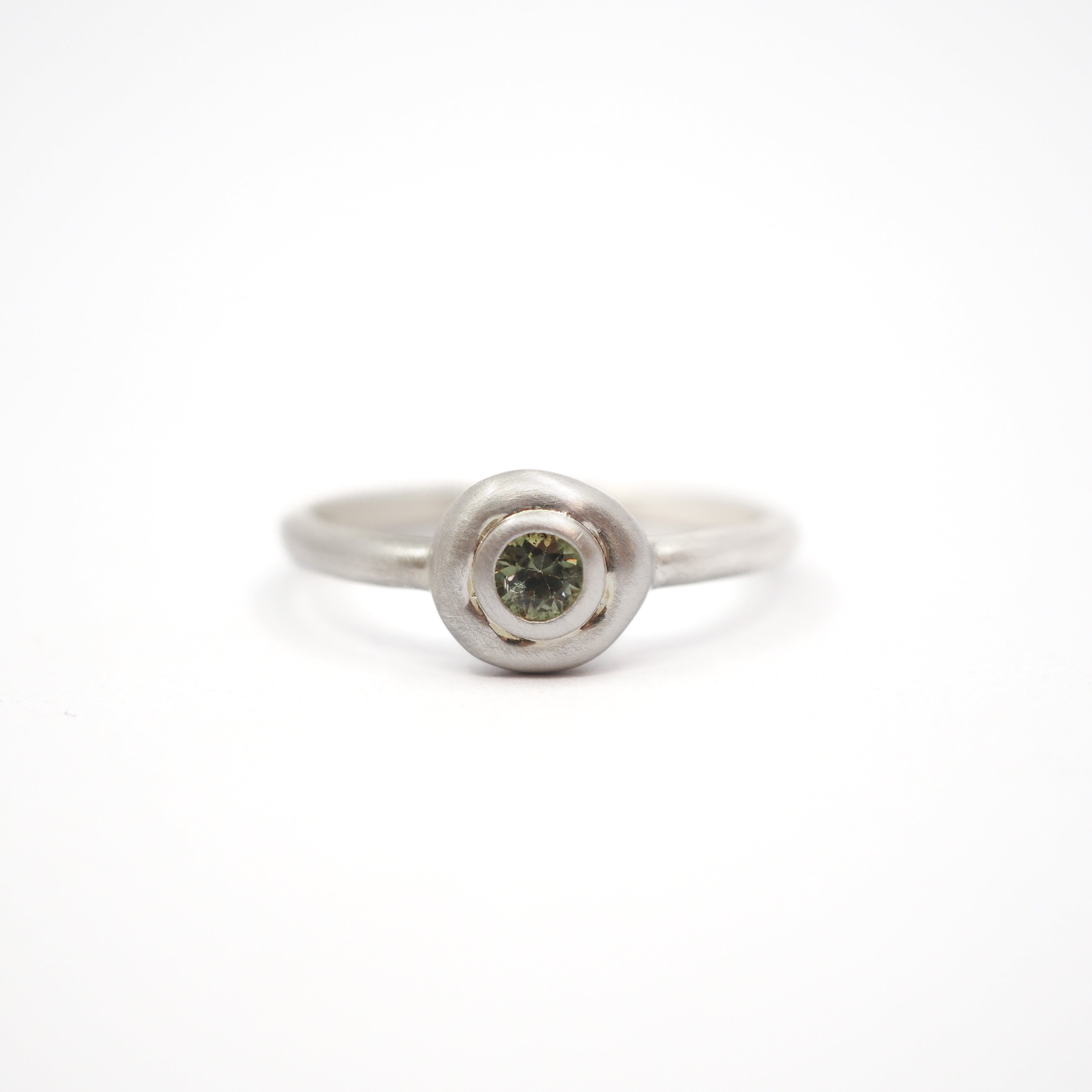 A silver ring with a textured, circular bezel setting holding a small round green gemstone, photographed on a seamless white background.