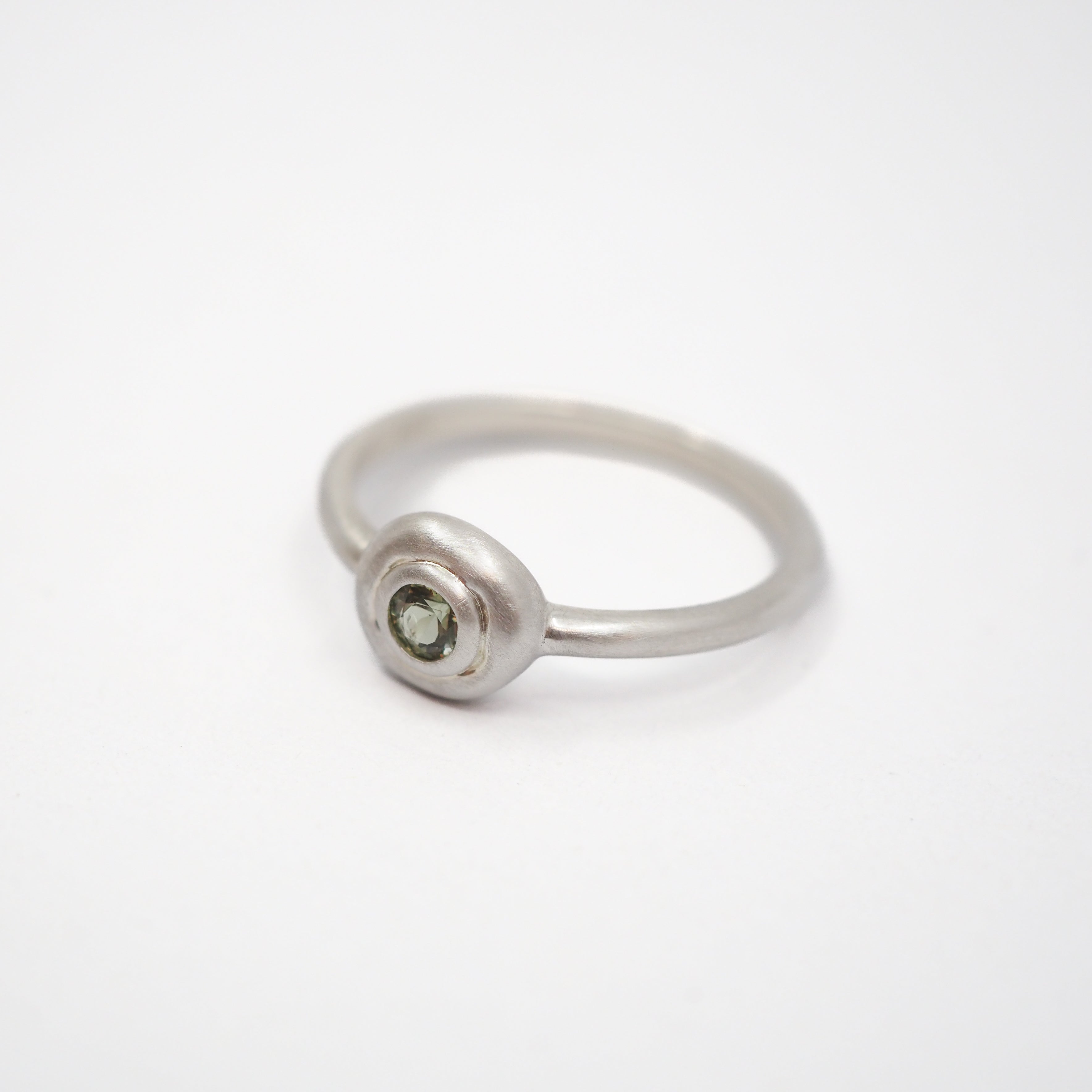A minimalist sterling silver ring featuring a small, bezel-set round green gemstone on a clean white background.