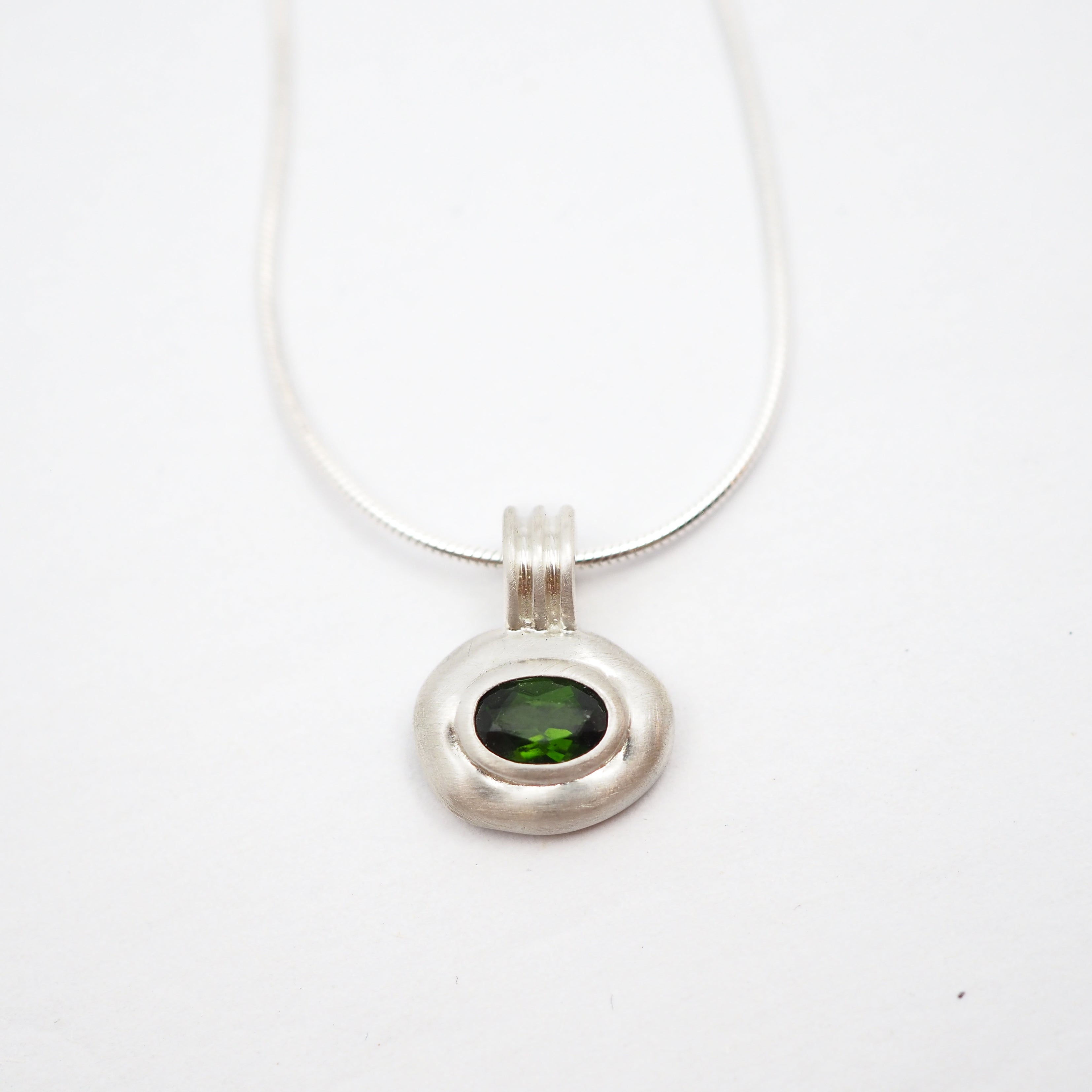 A silver pendant necklace featuring a textured, organic-shaped silver charm with a smooth, oval green gemstone in the center, suspended from a thin silver snake chain on a white background.