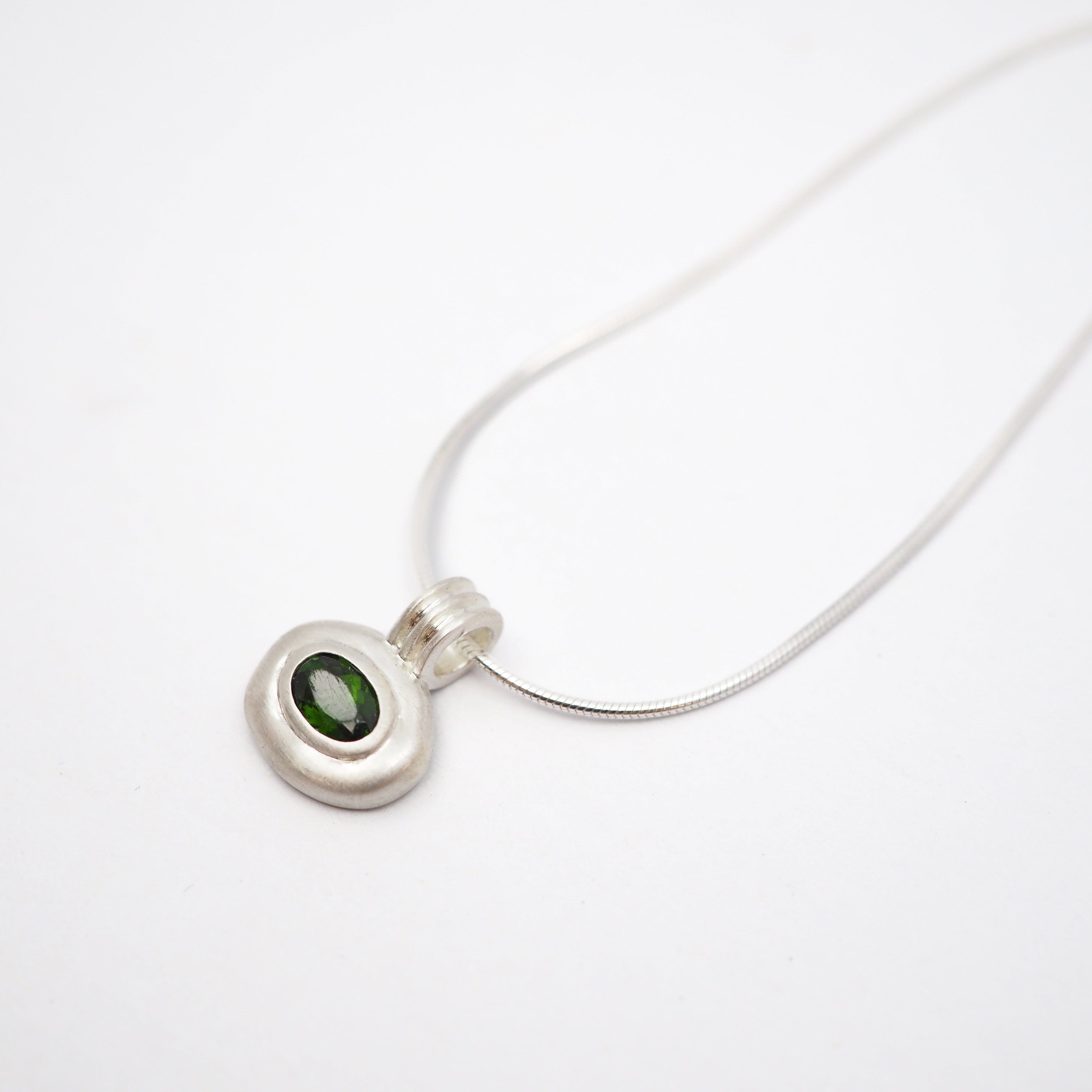 A silver pendant necklace featuring a textured, organic-shaped silver bezel holding a small oval green gemstone, suspended from a delicate silver snake chain on a plain white background.