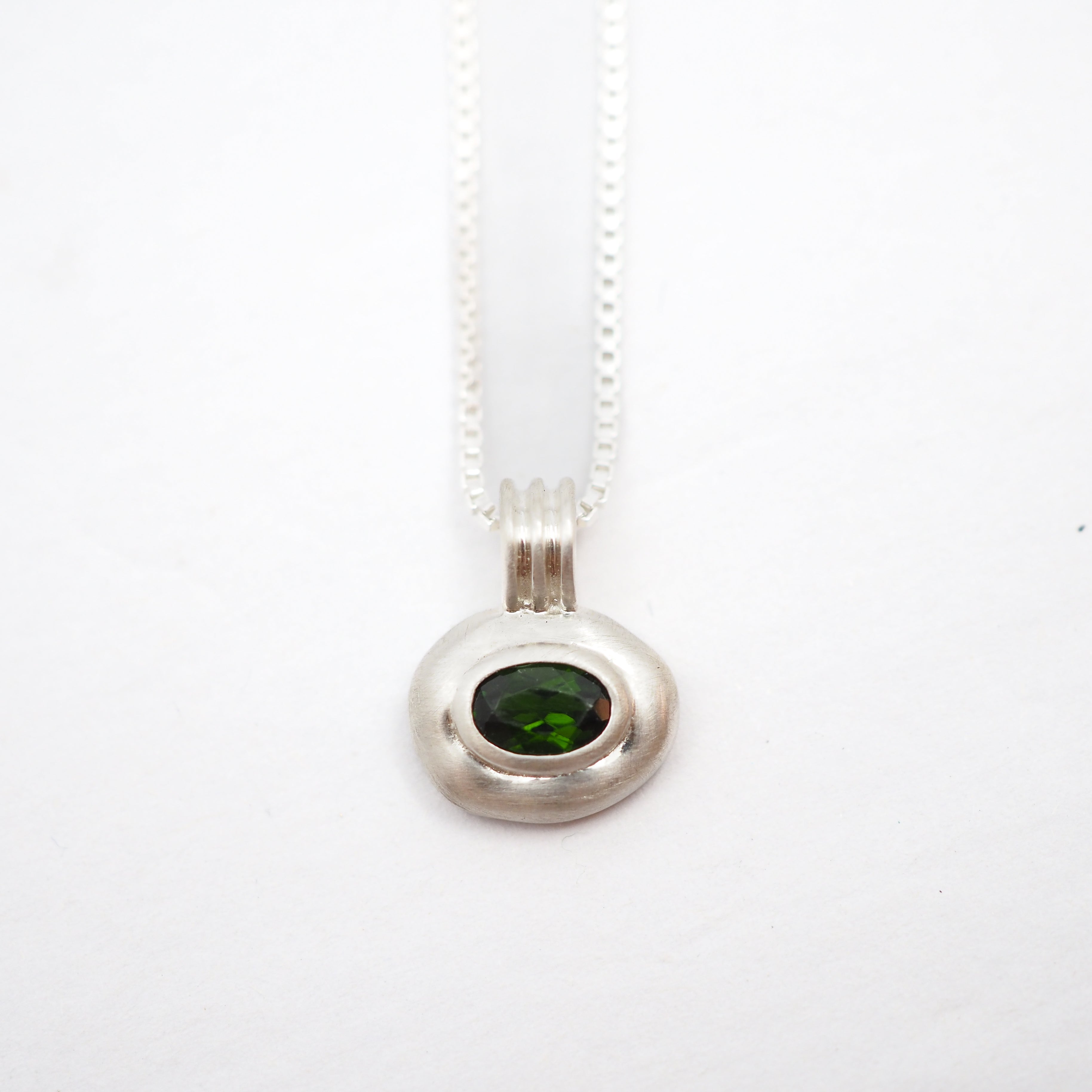 A silver pendant necklace featuring a bezel-set oval green gemstone on a delicate silver chain, photographed against a plain white background.