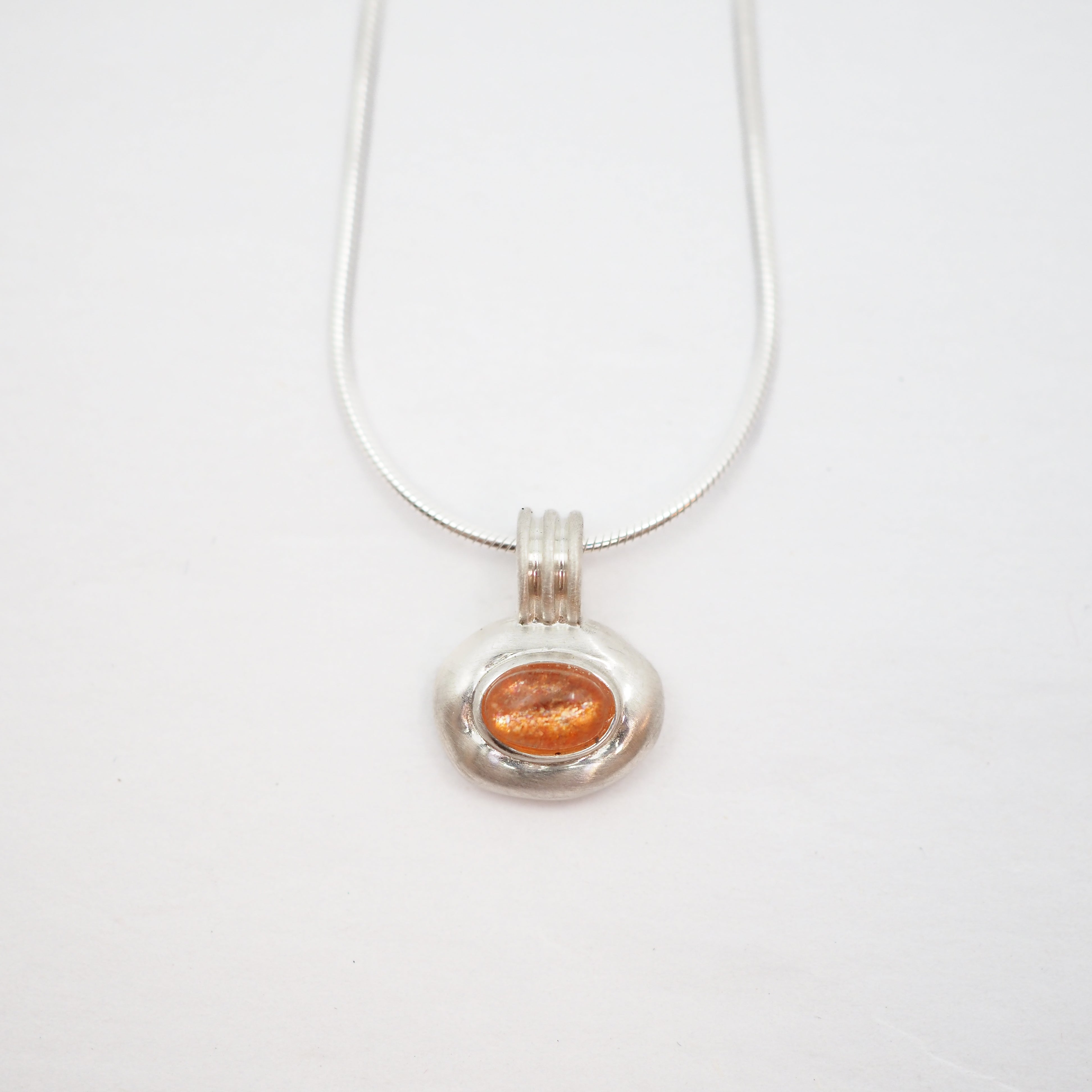 A silver pendant necklace featuring a textured, organic-shaped silver charm with a smooth, oval orange gemstone in the center, suspended from a thin silver snake chain on a white background.