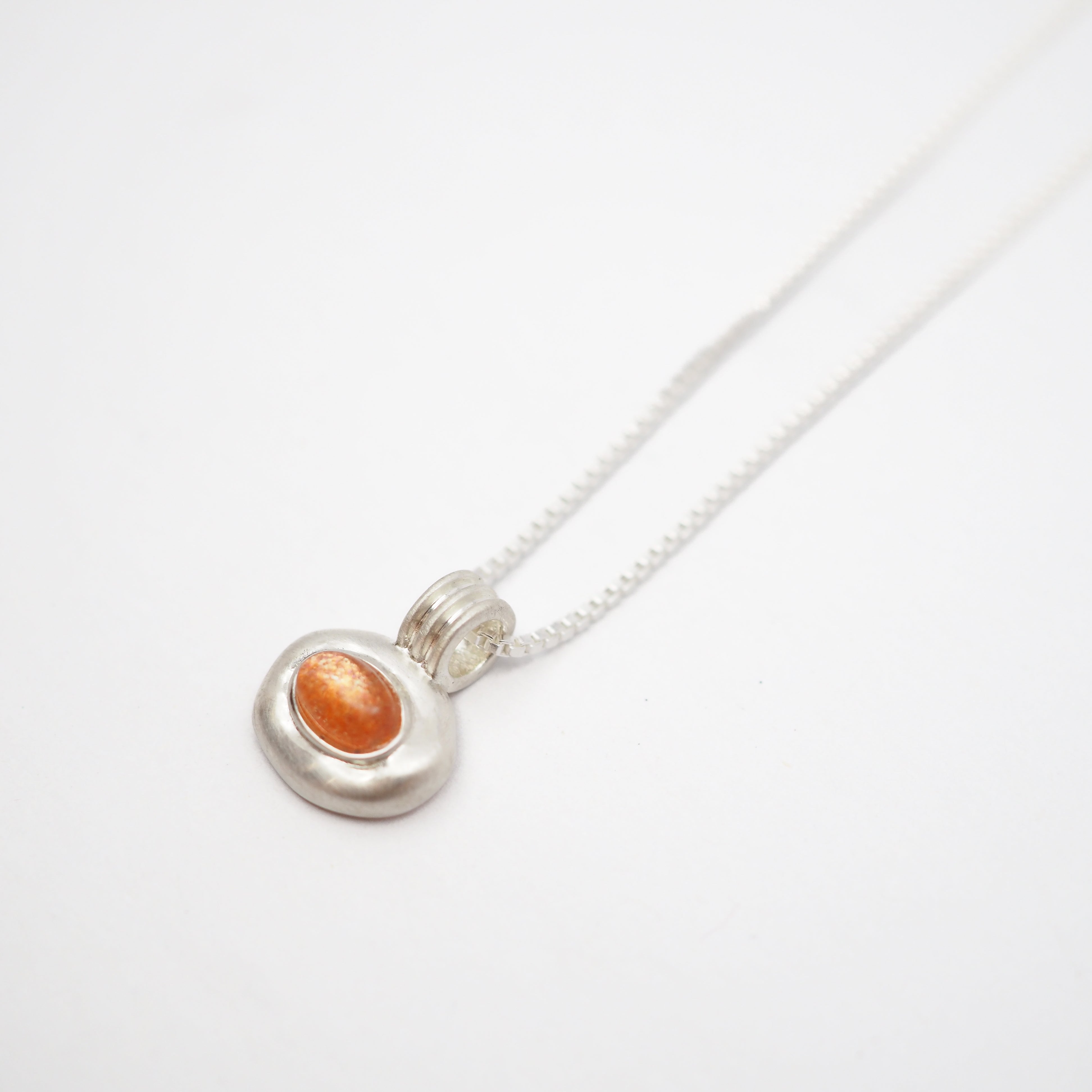 A silver pendant necklace featuring a textured, irregular silver oval setting with a smooth, polished orange cabochon gemstone, suspended on a delicate silver chain against a clean white background.