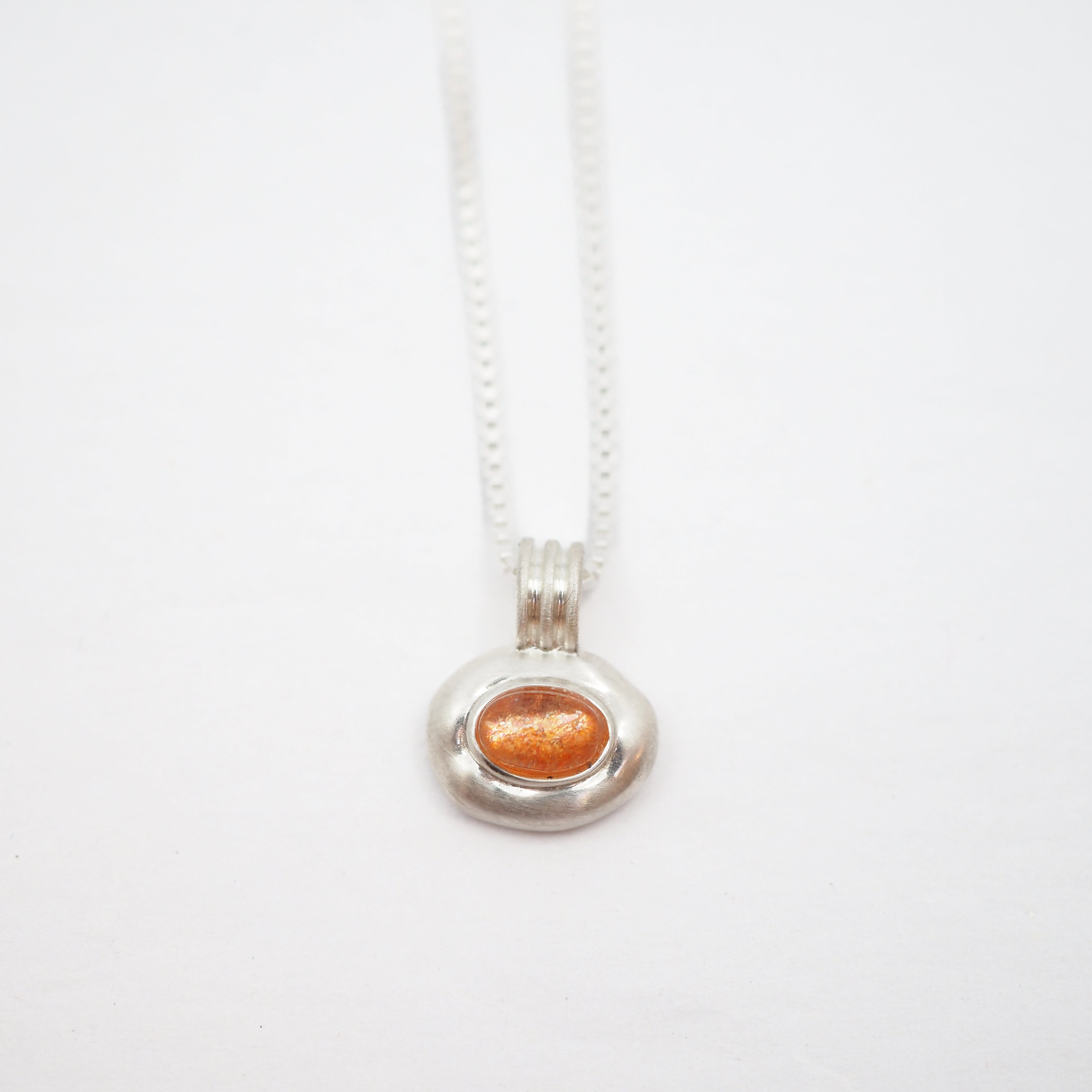 A silver chain necklace with a small, irregularly shaped silver pendant holding a textured, metallic orange gemstone, shown on a plain white background.