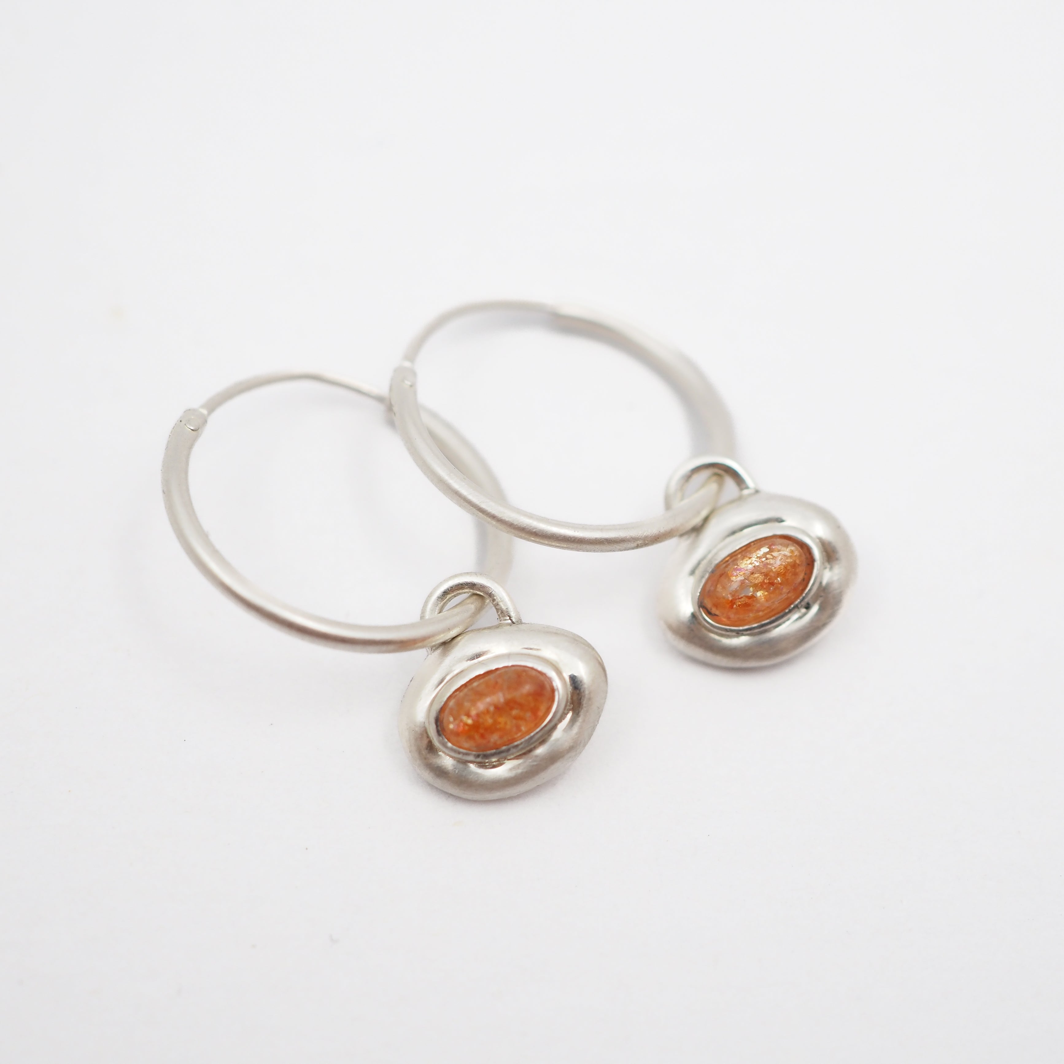 A pair of silver hoop earrings with small, bezel-set oval orange stones, displayed on a clean white background.