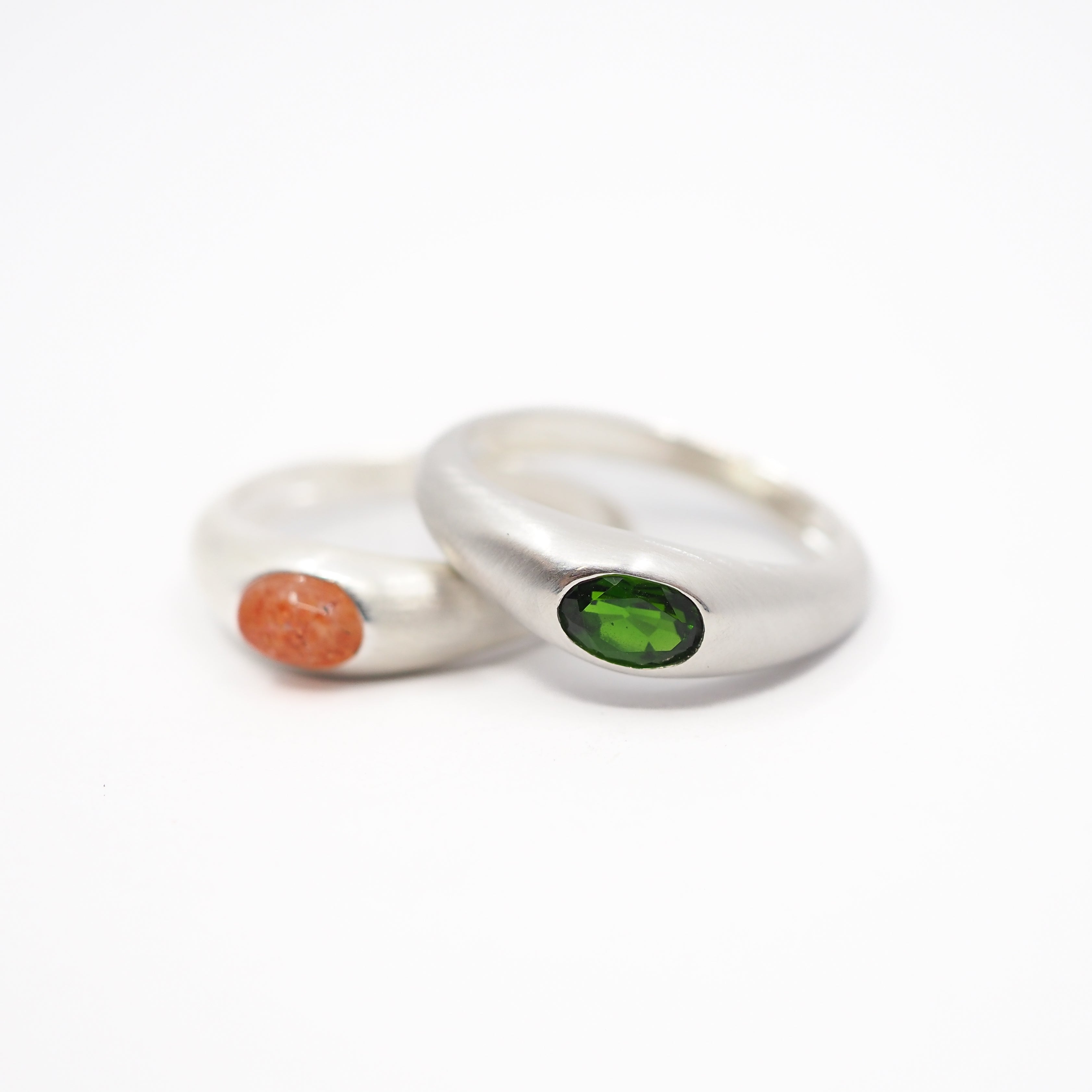 Two minimalist silver rings with oval inlays, one featuring a textured orange stone and the other a smooth green stone, displayed on a clean white background.
