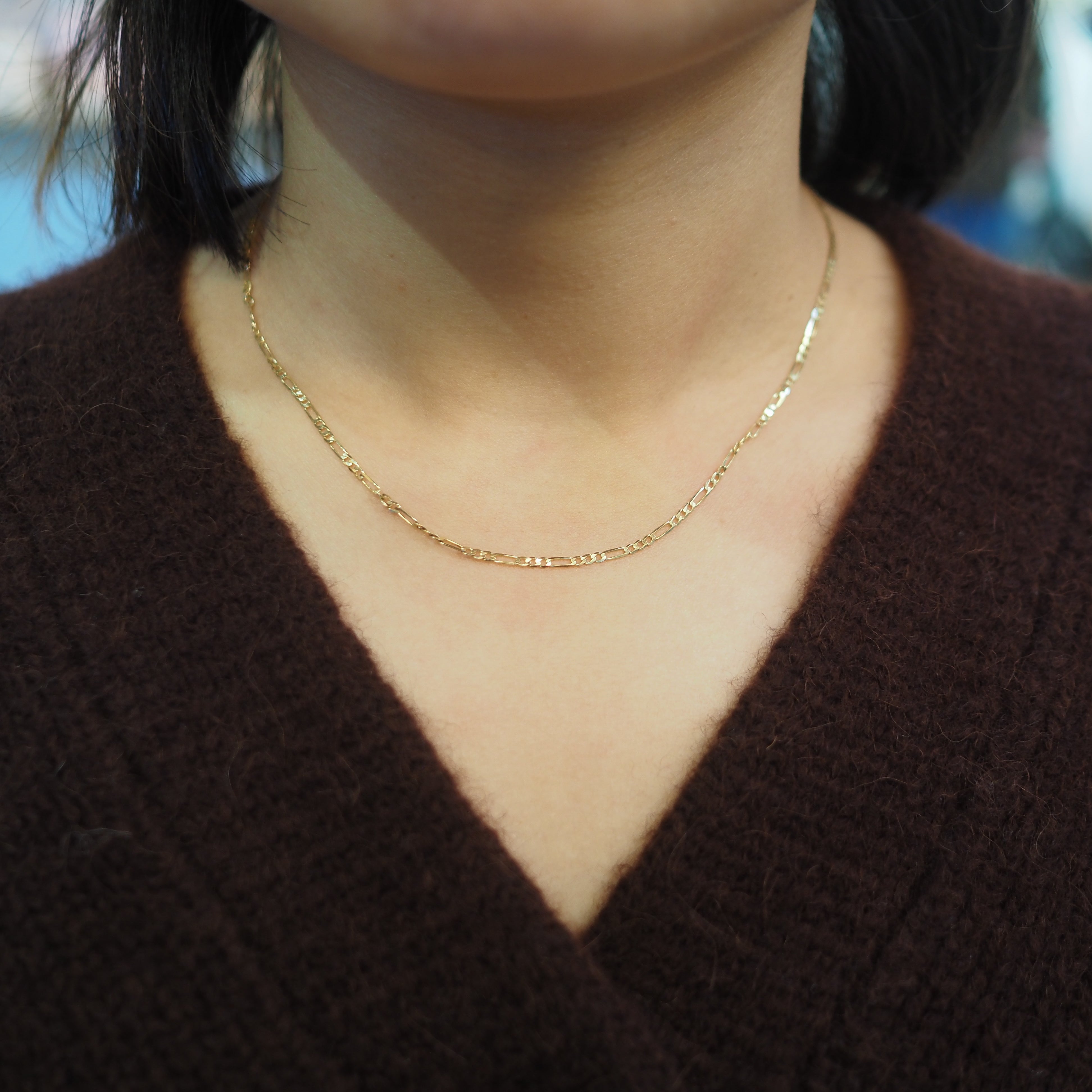 A close-up of a person wearing a delicate gold chain necklace styled over a dark brown V-neck sweater.