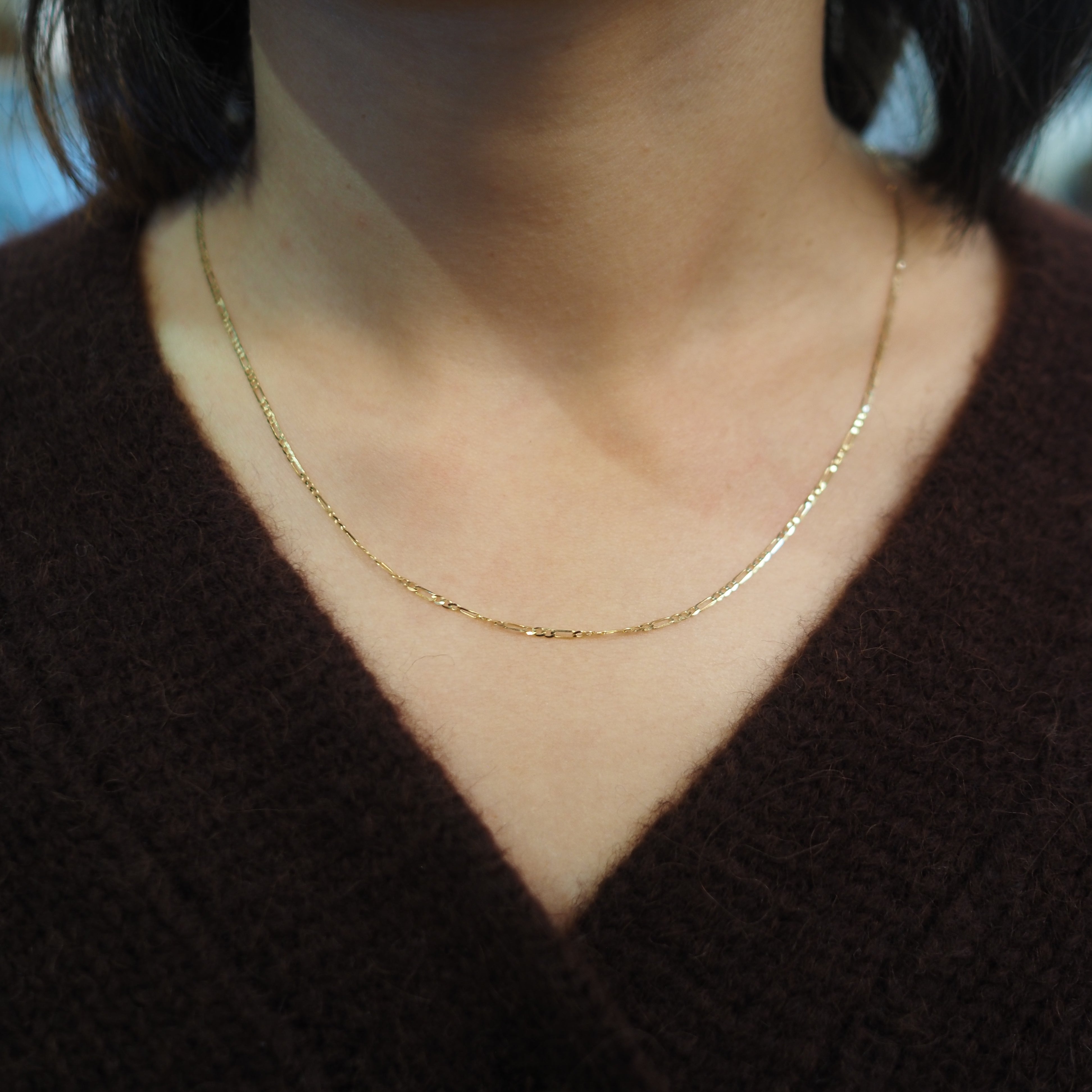 A close-up of a person's neck and chest wearing a delicate gold chain necklace over a dark brown V-neck sweater.