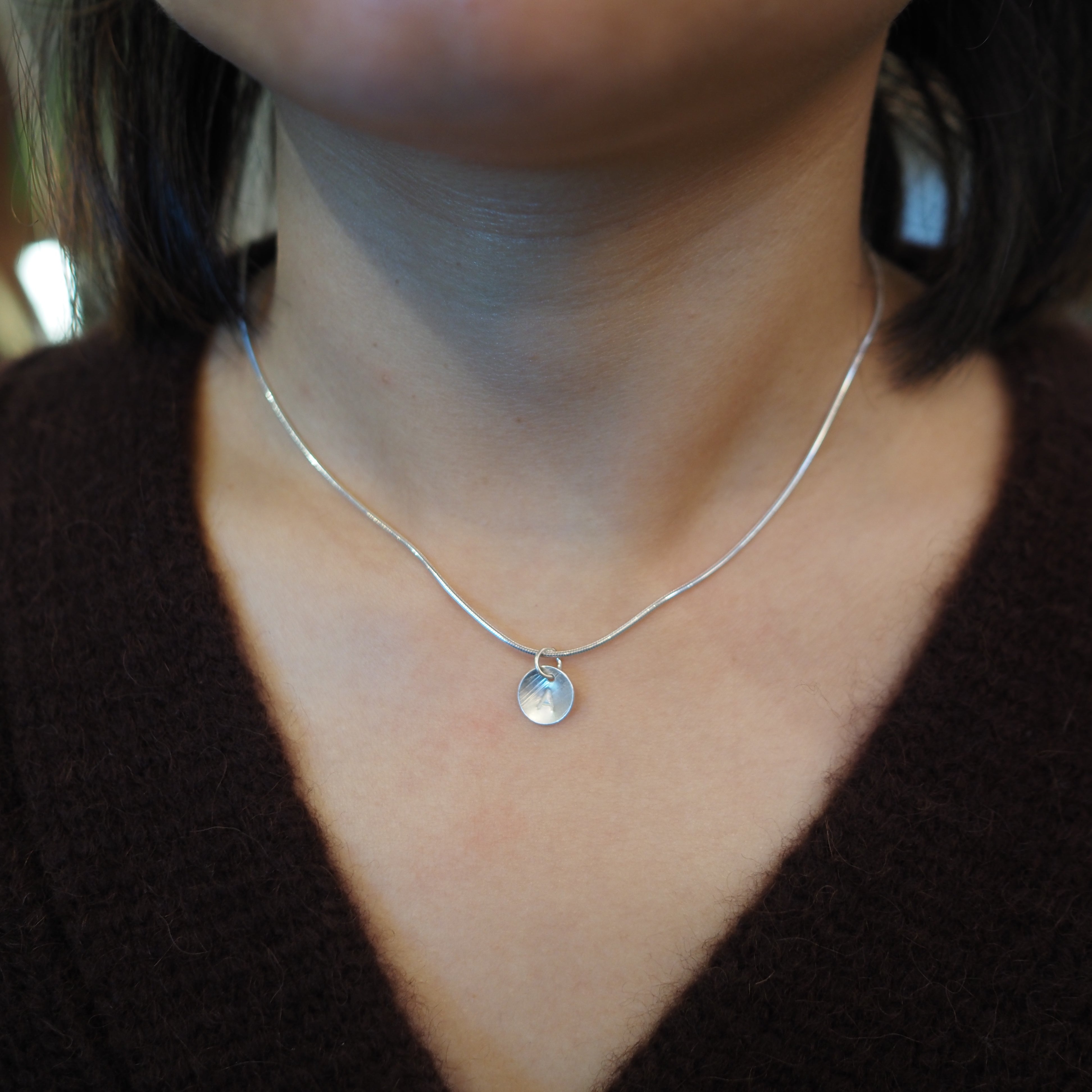 A close-up of a person's neck and chest wearing a delicate silver chain necklace with a small round silver pendant, paired with a dark brown knit top.
