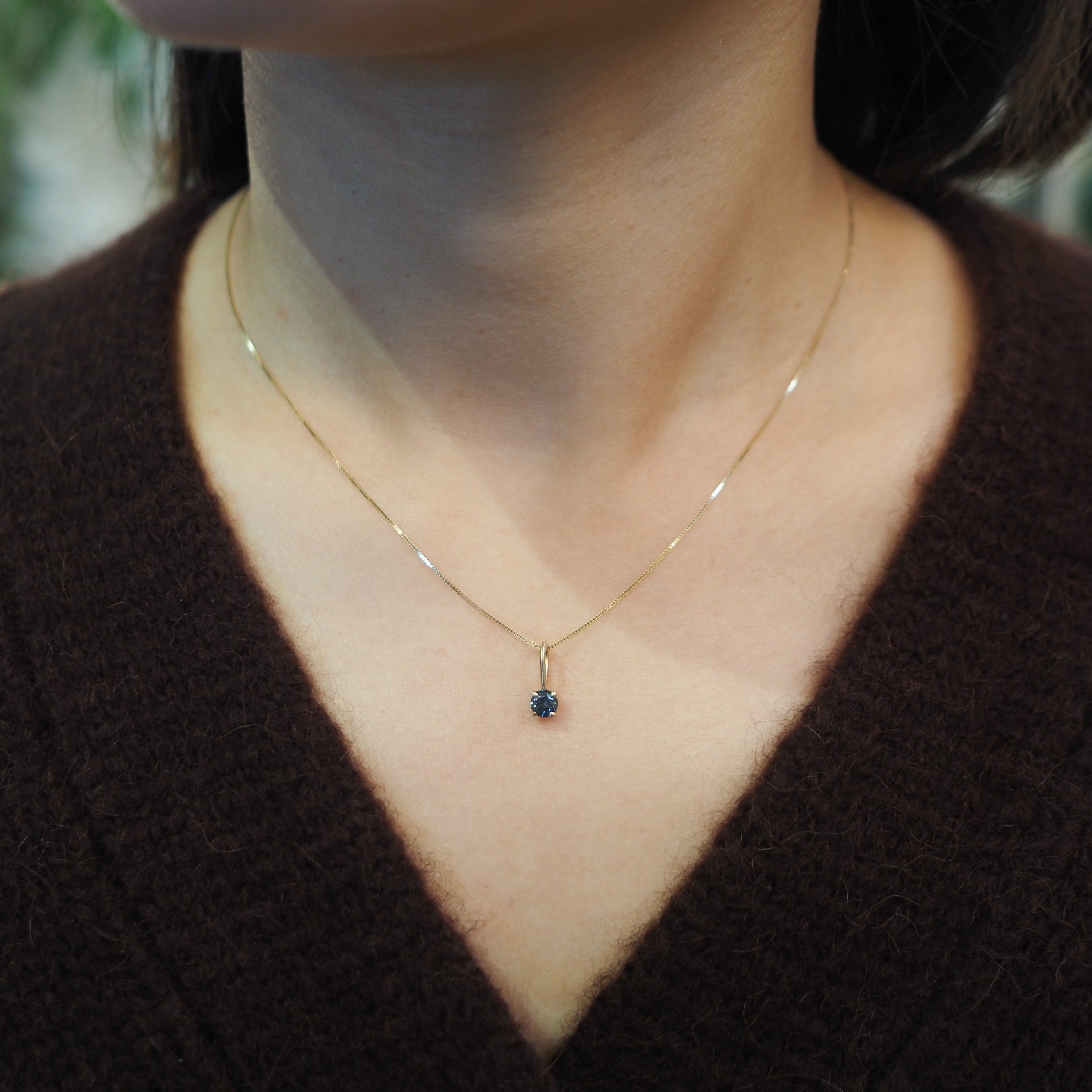 A close-up of a person wearing a delicate gold chain necklace with a small dark blue gemstone pendant, paired with a dark brown V-neck sweater.