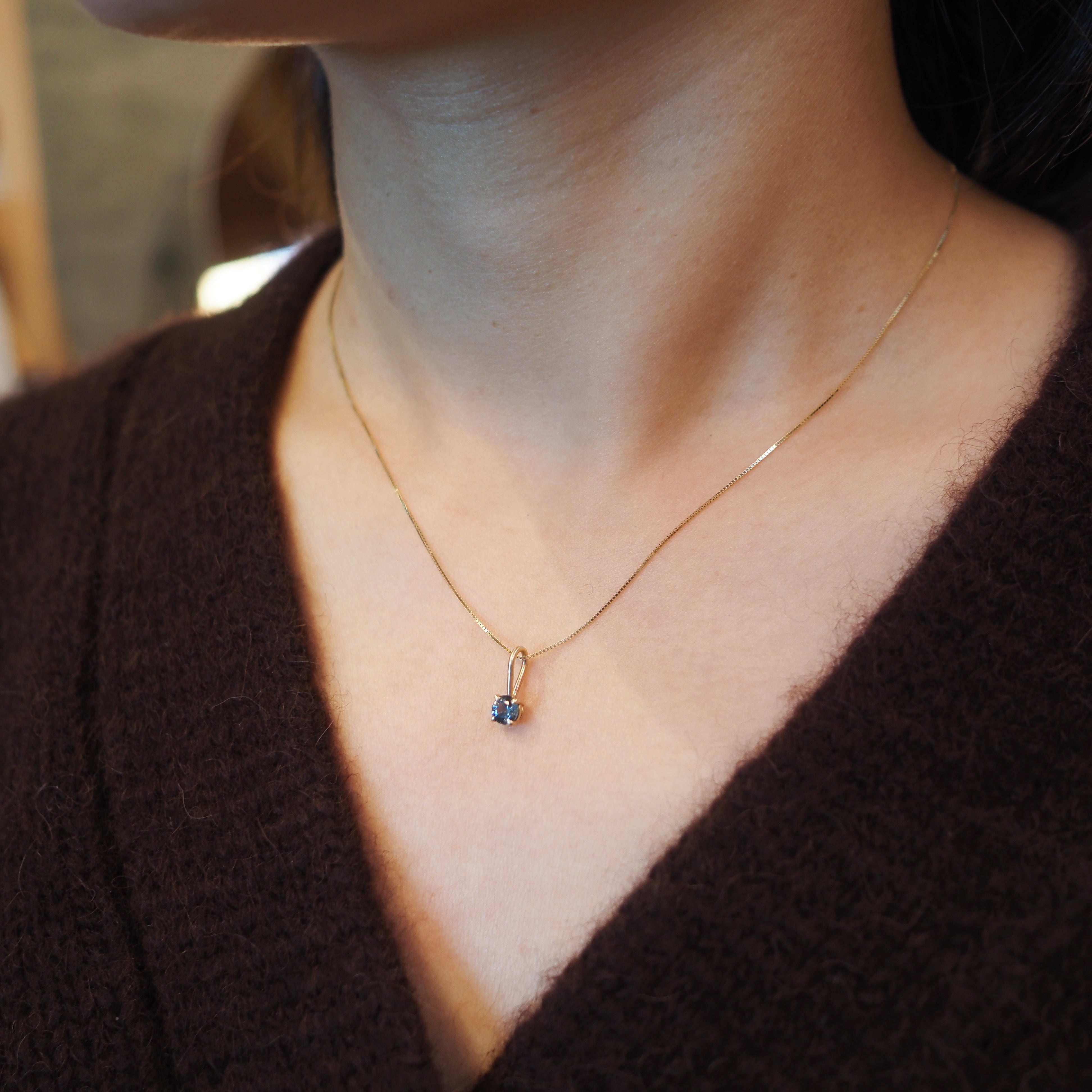 A close-up of a person's neck and chest wearing a delicate gold chain necklace with a small blue gemstone pendant, paired with a dark brown knit top.