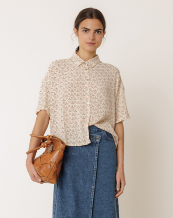 Woman wearing a patterned blouse and jeans, holding a brown leather bag on a plain background