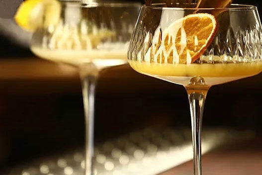 Two cocktail glasses with garnishes on a bar counter, blurred bottle in the background